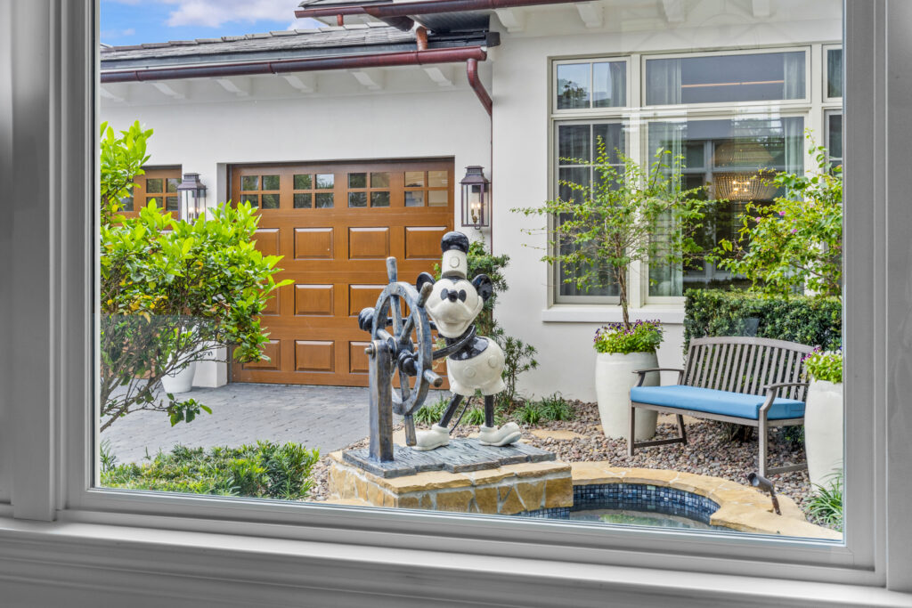 View through a window of a Disney-themed luxury vacation home courtyard featuring a Steamboat Willie Mickey Mouse statue at the helm, surrounded by lush landscaping, a bench, and a modern garage.
